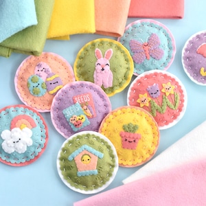 May include: Nine felt circles with embroidered spring-themed designs, including a bunny, a butterfly, an umbrella, a rainbow, a house, a flower, a plant, a seed packet, and an Easter egg. The circles are arranged on a light blue surface with pieces of felt in various colors in the background.