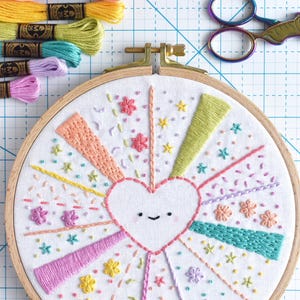 May include: A colorful embroidery hoop art piece featuring a smiling heart design with radiating lines and floral accents. The embroidery uses various pastel colors. Embroidery floss and scissors are also visible.