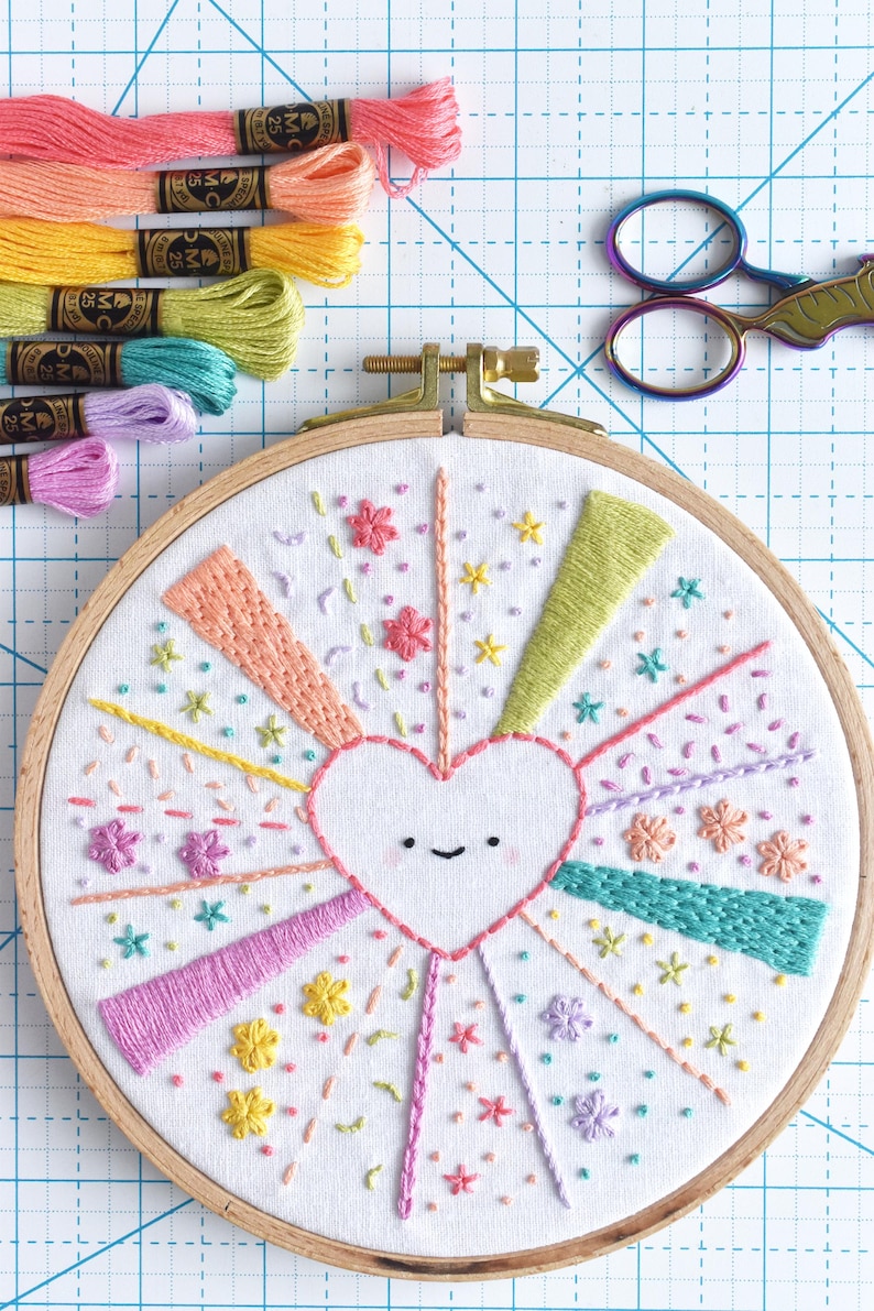 May include: A colorful embroidery hoop art piece featuring a smiling heart design with radiating lines and floral accents. The embroidery uses various pastel colors. Embroidery floss and scissors are also visible.