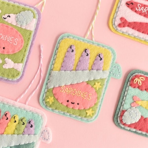 Può includere: Una collezione di ornamenti di sardine in feltro fatti a mano in vari colori. Ogni ornamento presenta un design diverso, tra cui sardine in scatola e singoli pesci, con la parola "Sardines" ricamata sopra. Gli ornamenti sono su uno sfondo rosa.