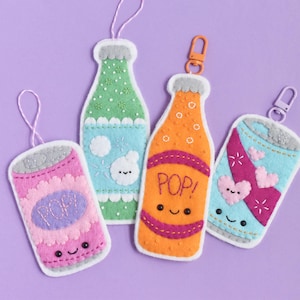 May include: Four felt ornaments shaped like beverage containers. Two are bottles and two are cans, featuring embroidered details, stitched faces, and the word POP. They include hanging loops or clips in pink, green, orange, and light blue colors.