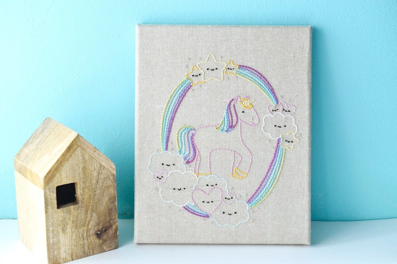 May include: A pink unicorn embroidered on a beige canvas with a rainbow and clouds. The unicorn has a gold crown and a rainbow mane and tail. The clouds have happy faces.