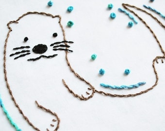 May include: Embroidered design of an otter swimming in blue water with brown outlines. The otter has black eyes, a black nose, and black whiskers. The water is represented by blue stitches and the otter's body is outlined in brown thread.