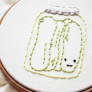 Pode incluir: Arte de bordado costurada à mão com um frasco de picles. O desenho está em tecido branco esticado dentro de um bastidor de bordado de madeira. Os picles e o contorno do frasco são em tons de verde e amarelo, com um rosto sorridente.