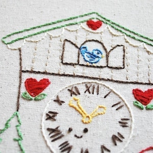 Puede incluir: Reloj de cuco bordado con un pájaro azul en la ventana, una esfera de reloj blanca con números romanos y corazones rojos con hojas verdes a cada lado.