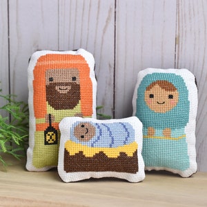May include: Three handmade felt Christmas ornaments depicting the Nativity scene. The figures are a brown-haired woman in a blue robe, a man with an orange beard in a yellow robe, and a baby swaddled in blue and yellow fabric in a brown manger.