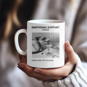 May include: White ceramic mug with a black and white image of a snake and cocktail glass. Text on the mug reads "EMOTIONAL SUPPORT SNAKE. SHEDS SKIN, NOT FEELINGS." The mug has a large, curved handle.