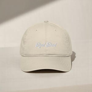 May include: A beige baseball cap with the embroidered text "Rad Dad" in a script font. The cap has a curved brim and a fabric strap closure. The hat is made of a soft, durable material.