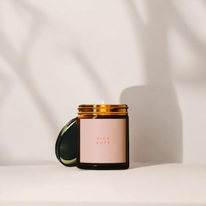 Puede incluir: Un tarro de vela de vidrio marrón con tapa dorada y una etiqueta rosa que dice "NICE BUTT" en texto rojo.