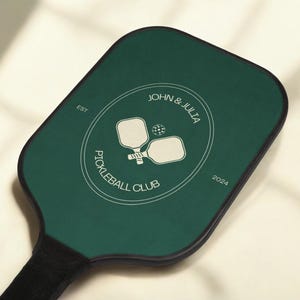Op de afbeelding: Een groene pickleball-paddle met een wit cirkelvormig logo met de tekst "John & Julia Pickleball Club" met de data "EST 2024".