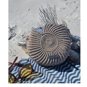May include: A beige macrame handbag with a spiral design, a gold chain strap, and a small "handmade" tag. The bag rests on a blue and white striped towel, with sunglasses and seashells nearby. The bag is approximately 10 inches in diameter.