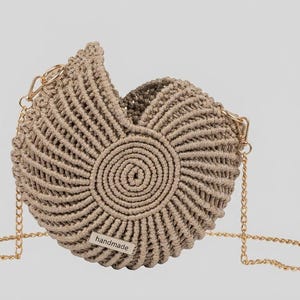 May include: A beige macrame handbag with a spiral design in the center. The bag has a gold chain strap and a small "handmade" tag. The bag's design resembles a seashell.