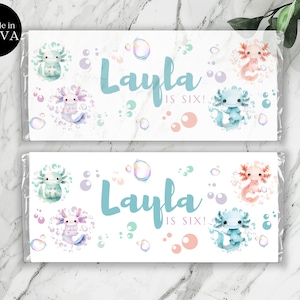 Puede incluir: Dos envoltorios de barra de chocolate imprimibles con un lindo diseño de ajolote. Los envoltorios son blancos con un fondo azul claro y presentan el texto "Layla tiene seis años!" en una fuente azul claro. Los ajolotes están rodeados de burbujas rosas, moradas y azules.