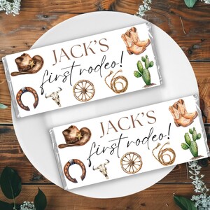 May include: Two white chocolate bar wrappers with a western theme. The wrappers feature the text "Jack's first rodeo!" with illustrations of cowboy hats, boots, a horseshoe, a skull, a wagon wheel, and a lasso.