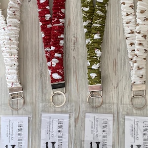 May include: Four fabric lanyards with metal key rings and clear plastic pouches. The lanyards feature festive patterns: white with snowmen, red with snowmen, olive green with snowmen, and white with reindeer. The text "CHENOWETH HAND" is visible.