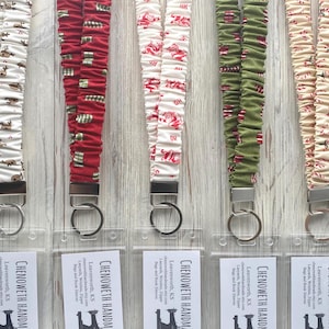 May include: Five fabric lanyards with gathered material, each featuring a silver metal clasp and a clear plastic ID badge holder. The patterns include reindeer on cream, gifts on red, Santa on cream, presents on green, and candy canes on tan.