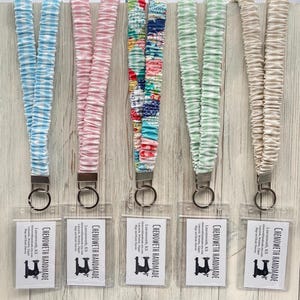 May include: Five fabric lanyards in various colors: light blue, pink, patterned, mint green, and cream. Each lanyard has a metal clasp and a clear plastic card holder. The lanyards are made by Chenoweth Handmade.