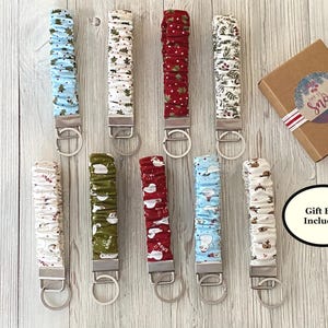 May include: A collection of fabric keychains in various holiday-themed patterns, including trees, snowmen, and berries. Each keychain has a silver-colored metal clasp and ring. A small gift box with a "Let it Snow" sticker is also included.