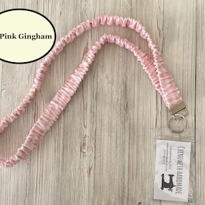 Puede incluir: Un cord&oacute;n rosa y blanco a cuadros gingham con un cierre y un llavero plateados. El cord&oacute;n tiene un soporte de pl&aacute;stico transparente con una tarjeta de visita. El texto "Icy Pink Gingham" est&aacute; en un &oacute;valo negro.