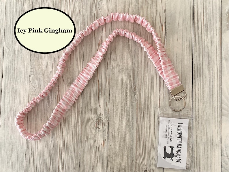 Puede incluir: Un cord&oacute;n rosa y blanco a cuadros gingham con un cierre y un llavero plateados. El cord&oacute;n tiene un soporte de pl&aacute;stico transparente con una tarjeta de visita. El texto "Icy Pink Gingham" est&aacute; en un &oacute;valo negro.