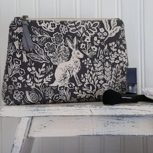 May include: A gray zippered pouch with a white floral and rabbit print. The pouch is sitting on a white wooden stool with makeup brushes and a compact.
