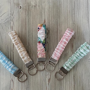 May include: Five fabric key fobs in various patterns and colors, including blue gingham, beige, pink gingham, and a patchwork design. Each fob has a metal key ring and a fabric loop. The key fobs are arranged on a light wood surface.