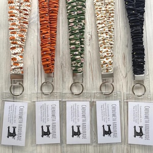May include: Five fabric lanyards with gathered, scrunchie-style straps in various patterns including pumpkins, orange, green, candy corn, and black. Each lanyard features a silver metal clasp and a clear plastic ID badge holder with a business card.