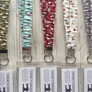 May include: Five fabric lanyards with gathered, ruffled designs in olive green, light blue, red, white, and gray, each featuring a holiday-themed print, a silver-toned metal clasp, a circular key ring, and a clear plastic ID badge holder with a card.