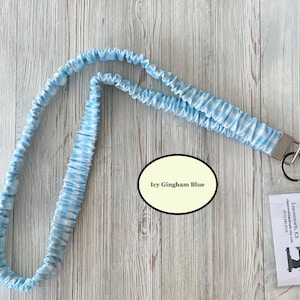 Puede incluir: Un cord&oacute;n con estampado de cuadros vichy azul claro y blanco con un cierre de metal y un llavero. El cord&oacute;n est&aacute; hecho de una tela suave y fruncida. El texto "Icy Gingham Blue" est&aacute; en un &oacute;valo. Una peque&ntilde;a etiqueta con texto es visible.