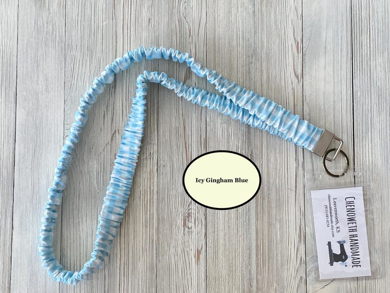 Puede incluir: Un cord&oacute;n con estampado de cuadros vichy azul claro y blanco con un cierre de metal y un llavero. El cord&oacute;n est&aacute; hecho de una tela suave y fruncida. El texto "Icy Gingham Blue" est&aacute; en un &oacute;valo. Una peque&ntilde;a etiqueta con texto es visible.
