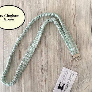 Puede incluir: Un cord&oacute;n de cuello verde hielo con estampado de cuadros vichy, con un llavero y cierre plateados. El cord&oacute;n est&aacute; hecho de tela fruncida verde claro y blanco. El texto "Icy Gingham Green" est&aacute; en un &oacute;valo negro.