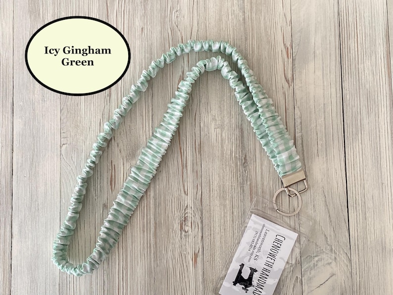 Puede incluir: Un cord&oacute;n de cuello verde hielo con estampado de cuadros vichy, con un llavero y cierre plateados. El cord&oacute;n est&aacute; hecho de tela fruncida verde claro y blanco. El texto "Icy Gingham Green" est&aacute; en un &oacute;valo negro.