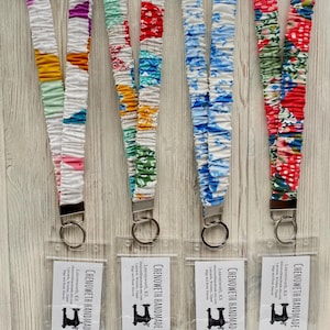 May include: A collection of fabric lanyards in various colors and patterns, including floral and patchwork designs. Each lanyard has a metal clasp and a clear plastic holder. The lanyards are displayed on a light wood-grain surface. The text "CHENOWETH HANDMADE" is visible.