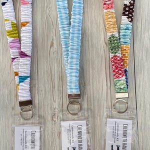 Puede incluir: Tres cordones de tela con cierres met&aacute;licos. Uno es blanco con formas geom&eacute;tricas coloridas, otro es de cuadros vichy azul claro, y el tercero es un patchwork de telas estampadas. Cada uno tiene una etiqueta transparente con texto.