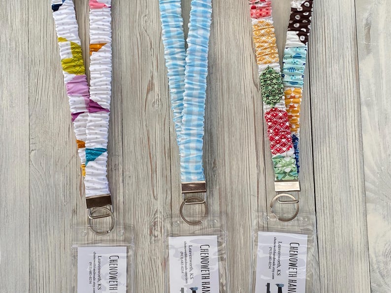 Puede incluir: Tres cordones de tela con cierres met&aacute;licos. Uno es blanco con formas geom&eacute;tricas coloridas, otro es de cuadros vichy azul claro, y el tercero es un patchwork de telas estampadas. Cada uno tiene una etiqueta transparente con texto.