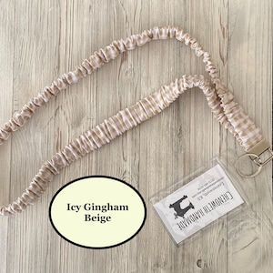 Puede incluir: Un cord&oacute;n con estampado de cuadros vichy beige y blanco con cierre met&aacute;lico y llavero. El texto "Icy Gingham Beige" est&aacute; en un &oacute;valo color crema. Tambi&eacute;n hay un tarjetero de pl&aacute;stico transparente con texto.