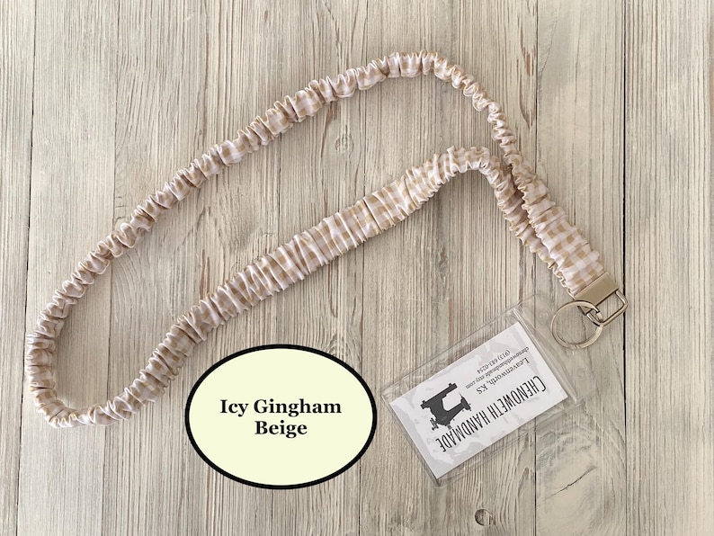 Puede incluir: Un cord&oacute;n con estampado de cuadros vichy beige y blanco con cierre met&aacute;lico y llavero. El texto "Icy Gingham Beige" est&aacute; en un &oacute;valo color crema. Tambi&eacute;n hay un tarjetero de pl&aacute;stico transparente con texto.