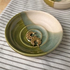 May include: A small, round ceramic dish in green, teal, and cream. Two gold-coloured hoop earrings with a decorative edge are inside the dish. The dish is on a striped fabric.