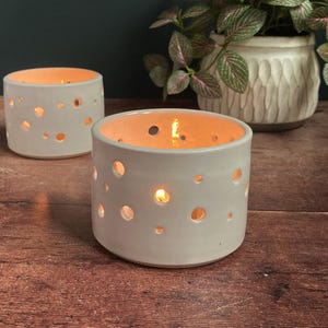 May include: Two white ceramic candle holders with a dotted pattern and a lit tea light inside each. The candle holders are on a wooden surface.