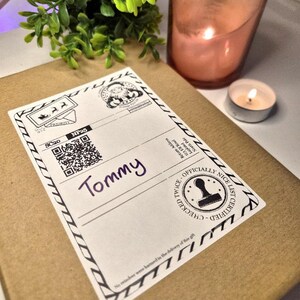 May include: A parcel with a label addressed to "Tommy." The label features a QR code, a Santa Claus graphic, and the text "OFFICIALLY NICE LIST CERTIFIED." A lit candle and a small plant are in the background.