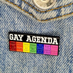 May include: A black enamel pin with the text "GAY AGENDA" in white letters. The pin is divided into seven sections, each representing a day of the week. Each section has a different colour and text, including "TACOS", "BE GAY", "SUPER GAY", and "BRUNCH GAY".