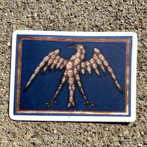 May include: A sticker featuring a blue background with a gold frame. The sticker depicts a stylised eagle made up of many human faces. The eagle has outstretched wings and is standing on two legs.