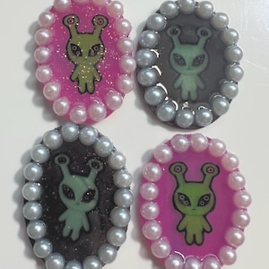 May include: Four oval-shaped alien-themed embellishments. Each features a green alien illustration on a pink or black background, surrounded by a border of faux pearls. The aliens have large eyes and antennae.
