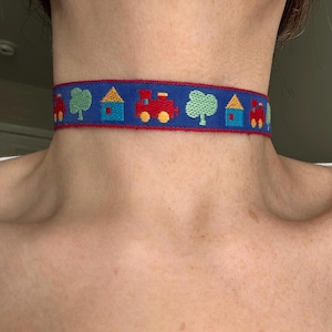 May include: A blue choker necklace with a repeating pattern of red trains, green trees, and blue houses with yellow roofs. The edges of the choker are trimmed with red. The necklace is worn around the neck.