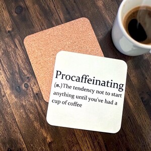 May include: A white square coaster with the word "Procaffeinating" and its definition printed on it. A second cork coaster is next to it. A white mug of coffee sits nearby on a wooden surface.