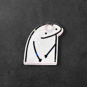 May include: A white sticker with a cartoon character design, featuring a sun-like emblem on its head. The figure has black lines for facial features and limbs, and the sticker has a holographic, glittery finish. The background is dark grey.