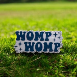 May include: A white enamel pin with blue lettering that reads "WOMP WOMP" with white stars on either side of the text.