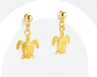 Op de afbeelding: Gouden oorbellen met hangende schildpadden op een witte displaystandaard met een houten basis.