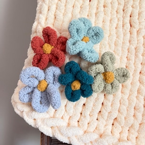 May include: A cream-coloured, chunky knit blanket decorated with five plush, flower-shaped appliqués. The flowers are in shades of red, blue, teal, and green, each with a yellow centre. The blanket is folded over a wooden ladder.