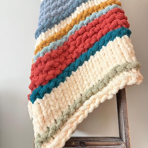 May include: A chunky knit throw blanket featuring diagonal stripes in cream, dusty blue, mustard yellow, terracotta red, teal, and sage green. The blanket is crafted from thick, soft yarn with a prominent, textured loop stitch pattern throughout.